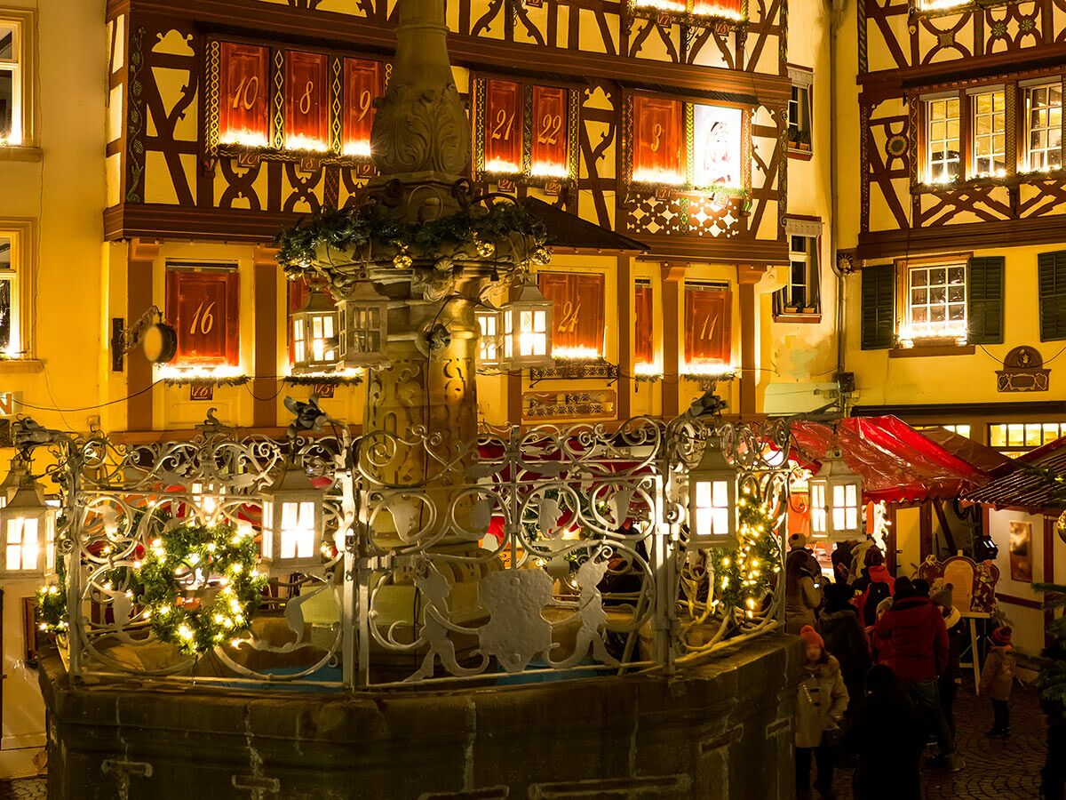 Festlicher Weihnachtsmarkt bei Nacht mit beleuchtetem Adventskalender-Gebäude, Brunnen mit Lichtern, geschmückten Ständen und Besuchern.