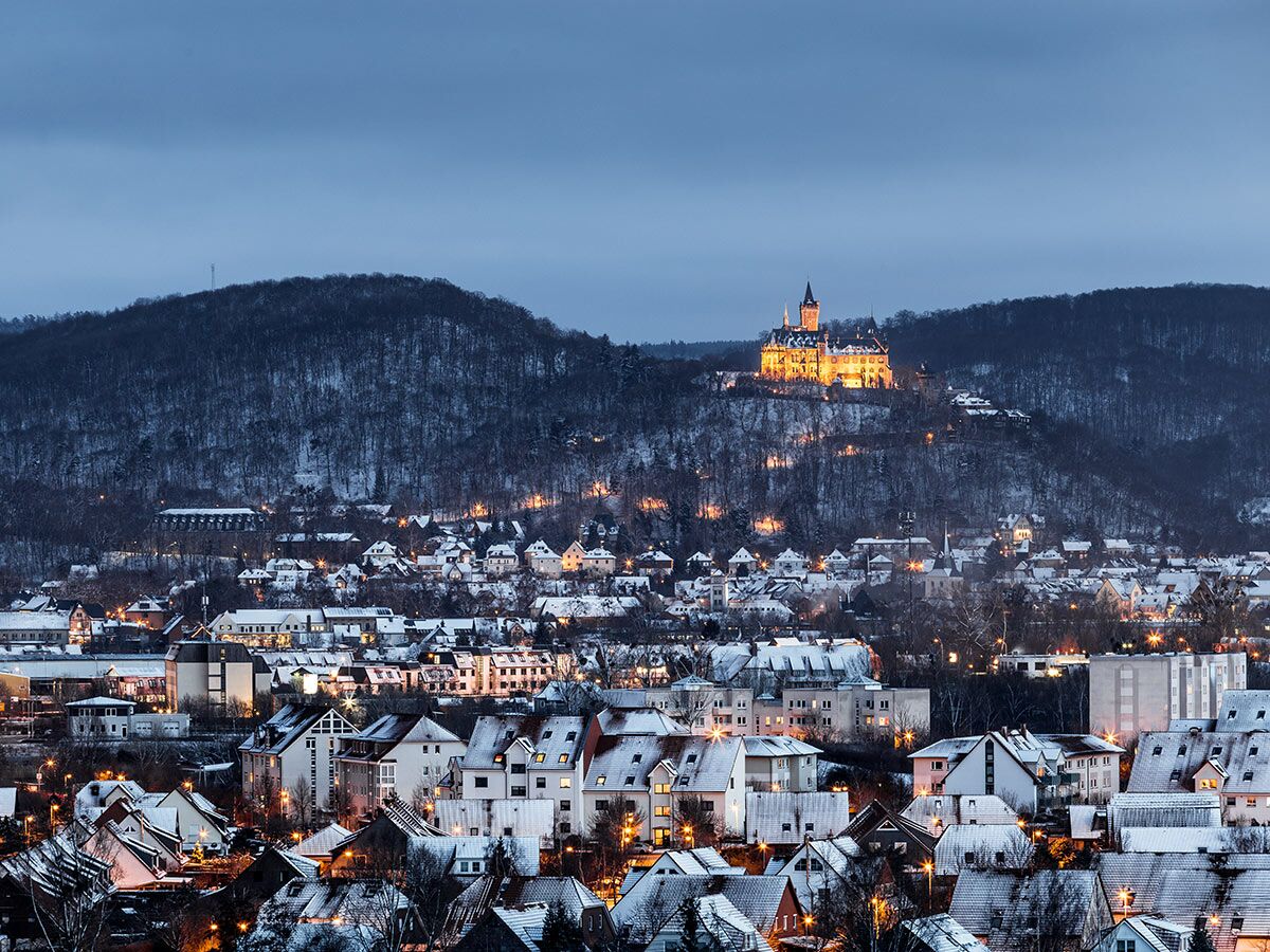 Wernigerode Schloss leuchtet goldgelb auf verschneitem Hügel über der winterlichen Stadt im Abendlicht, umgeben von dunklen Wäldern.
