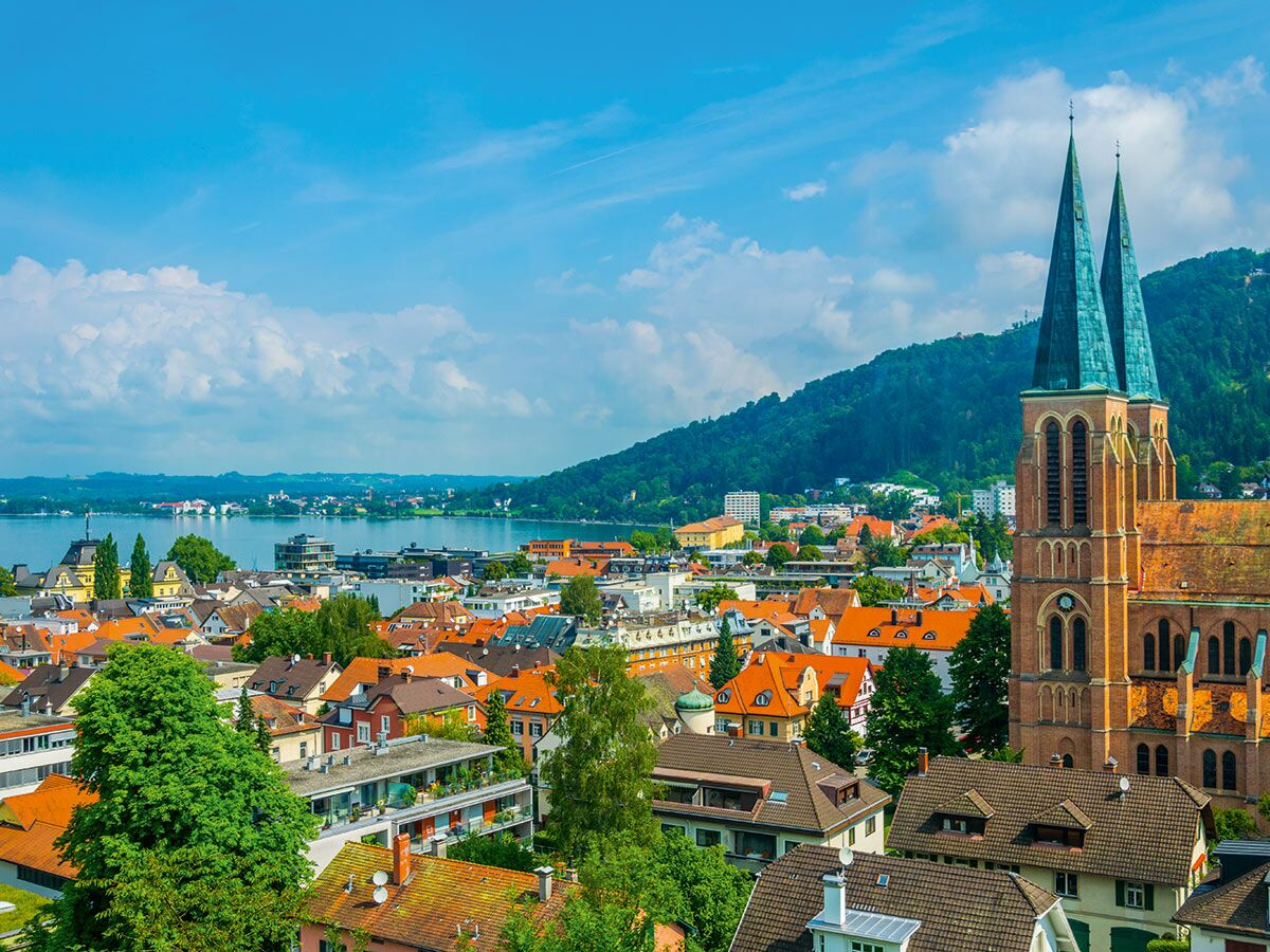 Panoramablick auf Bregenz, Österreich, mit der Evangelischen Kirche, roten Dächern, Bodensee und Pfänderberg unter blauem Himmel.
