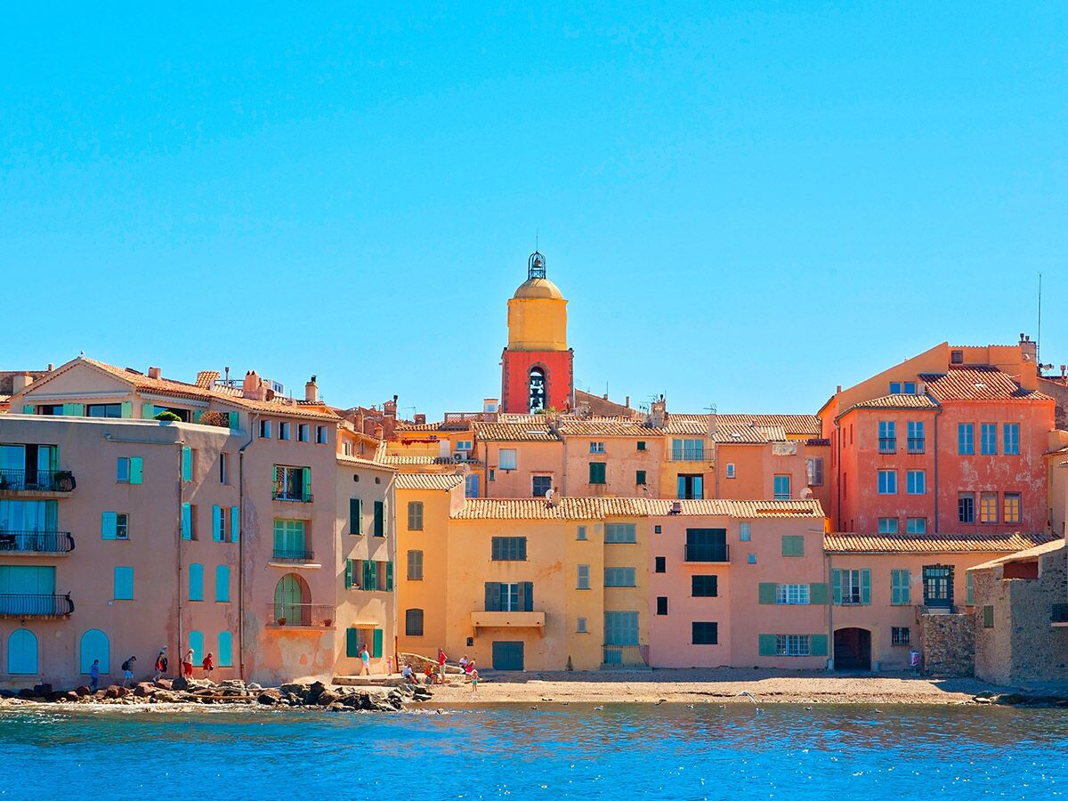 Malerische Küstenstadt St. Tropez mit farbenfrohen Häusern, rotem Glockenturm, blauem Meer und Menschen am Strand unter blauem Himmel.