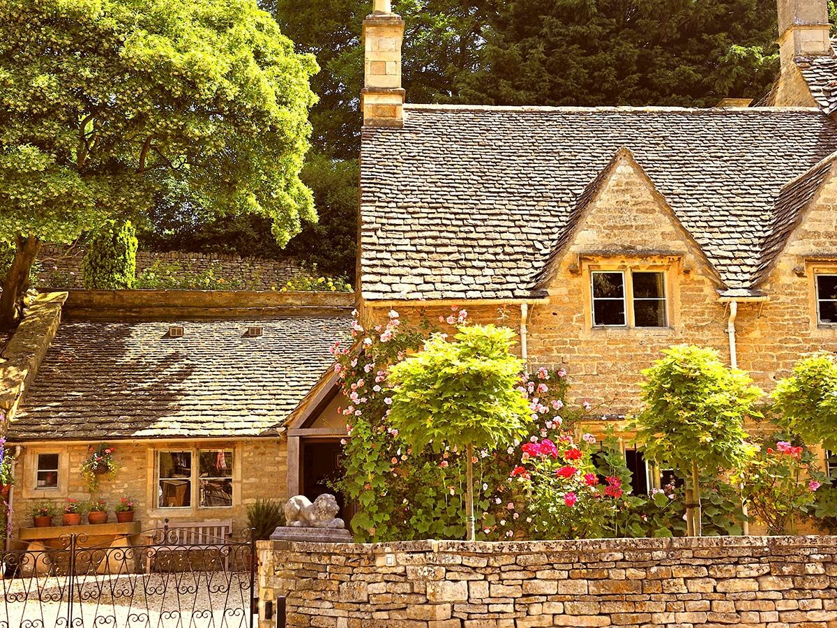 Malerisches Dorfbild mit traditionellen Steinhäusern, blühenden Gärten, Dachschindeln und hohen Bäumen in den englischen Cotswolds.