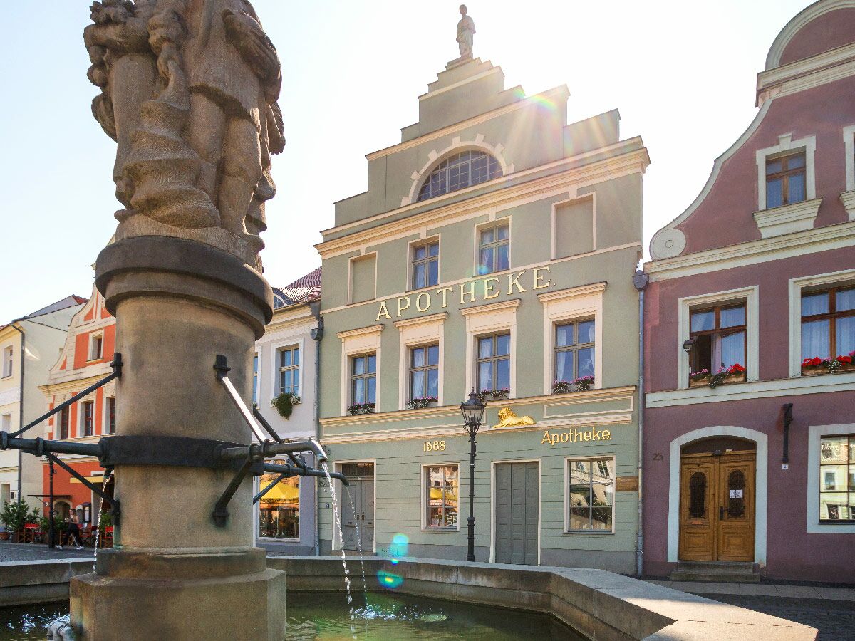 Historische Apotheke mit Stufengiebel und Brunnen auf einem sonnigen Marktplatz in einer europäischen Altstadt.