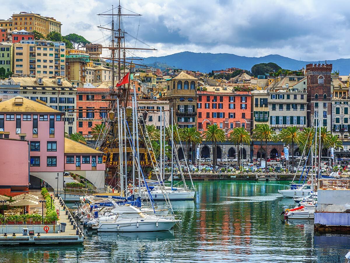 Vibranter Hafen in Genua, Italien, mit bunten historischen Gebäuden, einem Großsegler, modernen Segelbooten und Palmen am Wasser.