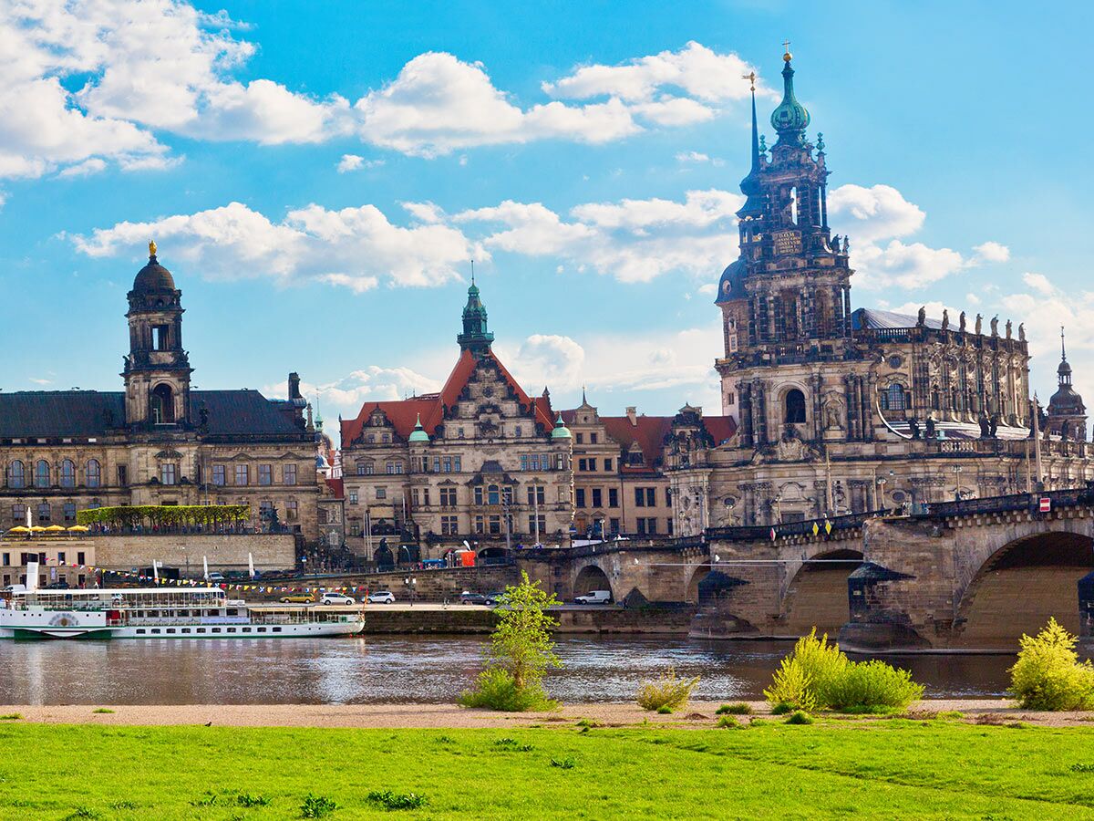 Historische Gebäude und Augustusbrücke an der Elbe in Dresden mit Flusskreuzfahrtschiff und grünem Ufer unter blauem Himmel.