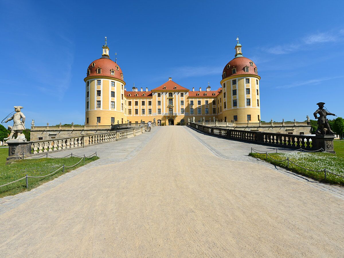 Schloss Moritzburg, ein gelbes Barockschloss mit roten Kuppeln und Zufahrtsweg, flankiert von Statuen unter blauem Himmel.