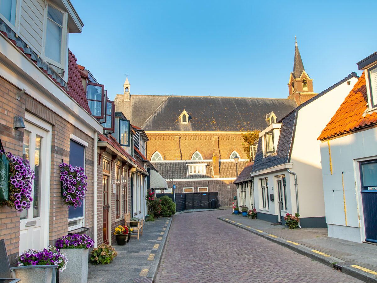 Ruhige Dorfstraße mit traditionellen Häusern, roten Dächern, lila Blumenampeln und einer Kirche unter blauem Himmel.