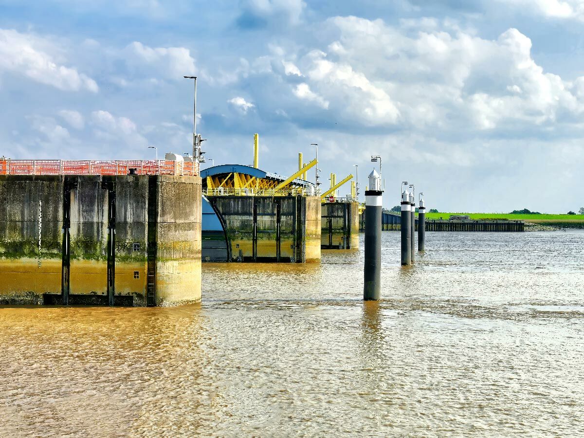 Große Schleusenanlage mit grauen Betonpfeilern, gelben Metallkonstruktionen und braunem Flusswasser unter bewölktem Himmel.