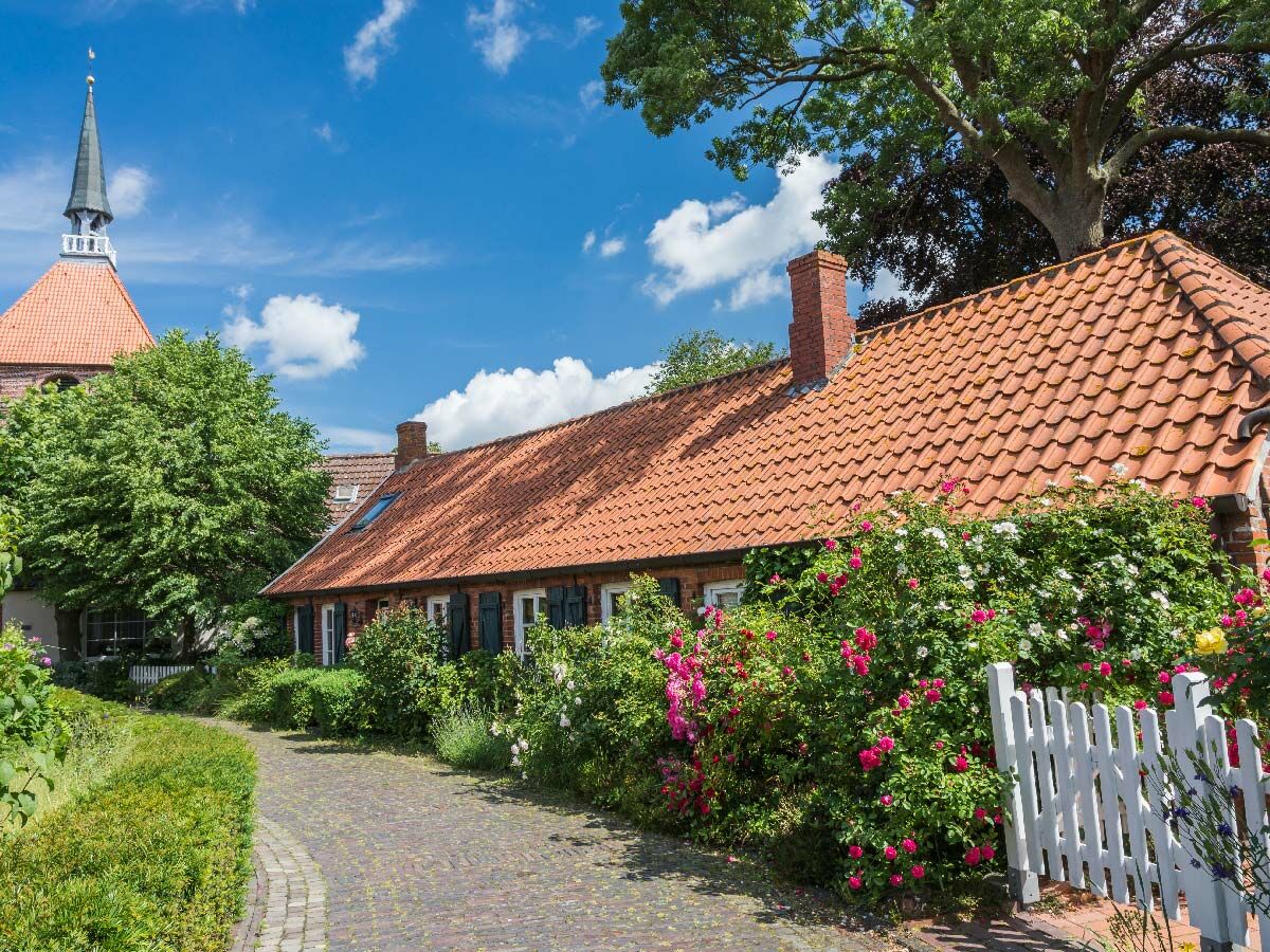 Historische Dorfgasse mit alten Häusern, roten Ziegeldächern, blühenden Rosen, weißem Zaun und Kirchturm unter blauem Himmel.