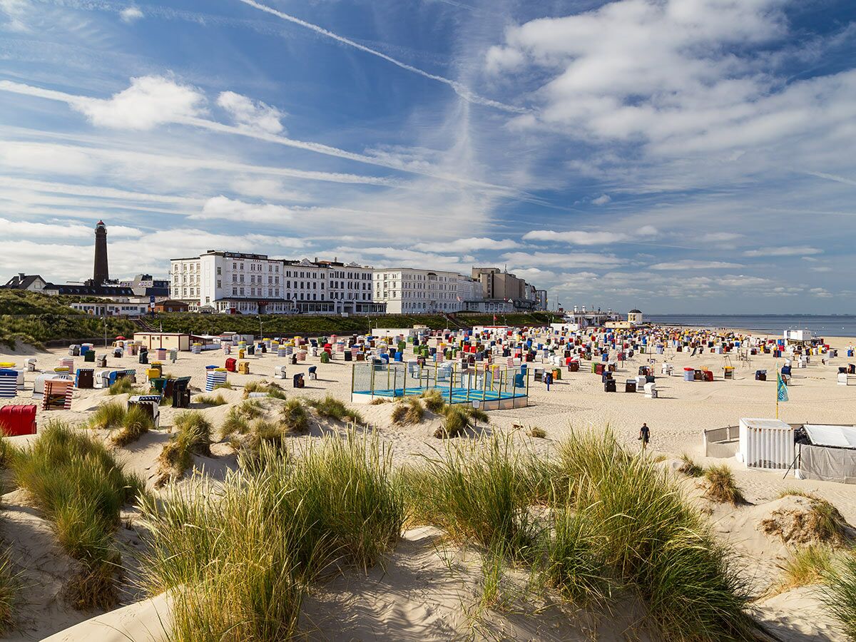 Breiter Sandstrand auf Langeoog mit bunten Strandkörben, Dünen, Leuchtturm und weißen Hotels unter blauem Himmel.
