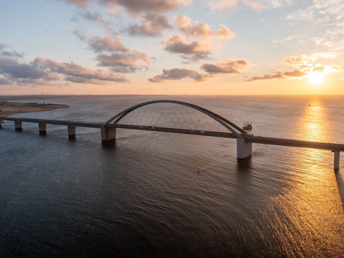 Die Fehmarnsundbrücke im Abendlicht, eine Bogenbrücke über das Meer mit Autos und Windrädern am Horizont.