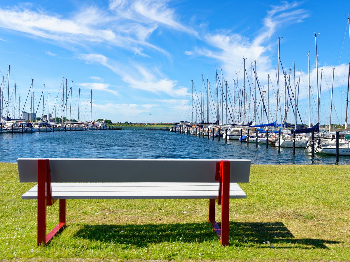 Weiße und rote Parkbank auf grünem Gras mit Blick auf einen sonnigen Yachthafen voller Segelboote unter blauem Himmel mit weißen Wolken.