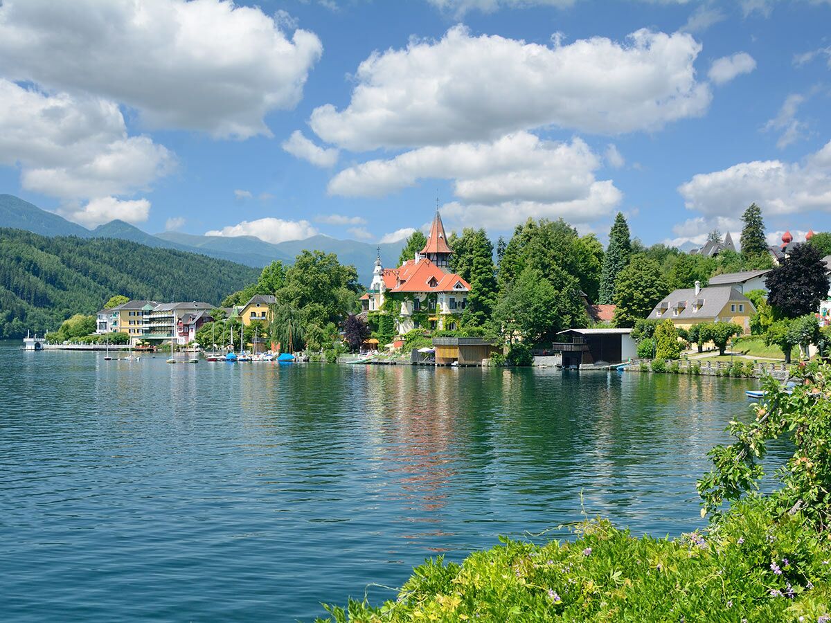 Idyllische Landschaft am Seeufer mit farbenfrohen Häusern, Booten und bewaldeten Bergen unter blauem Himmel mit Wolken.