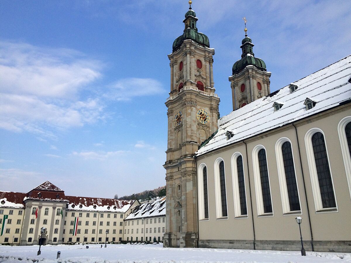 Die barocke Stiftskirche St. Gallen mit zwei Türmen und umliegenden Gebäuden, alles bedeckt mit frischem Schnee unter blauem Himmel.