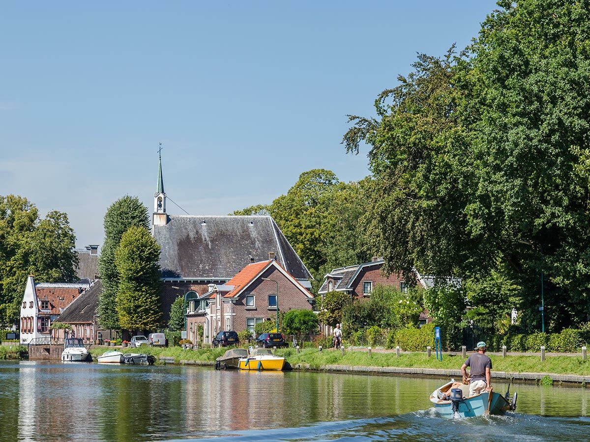 Mann fährt Boot auf Dorfkanal mit Kirche, Häusern und Bäumen im Hintergrund unter blauem Himmel.