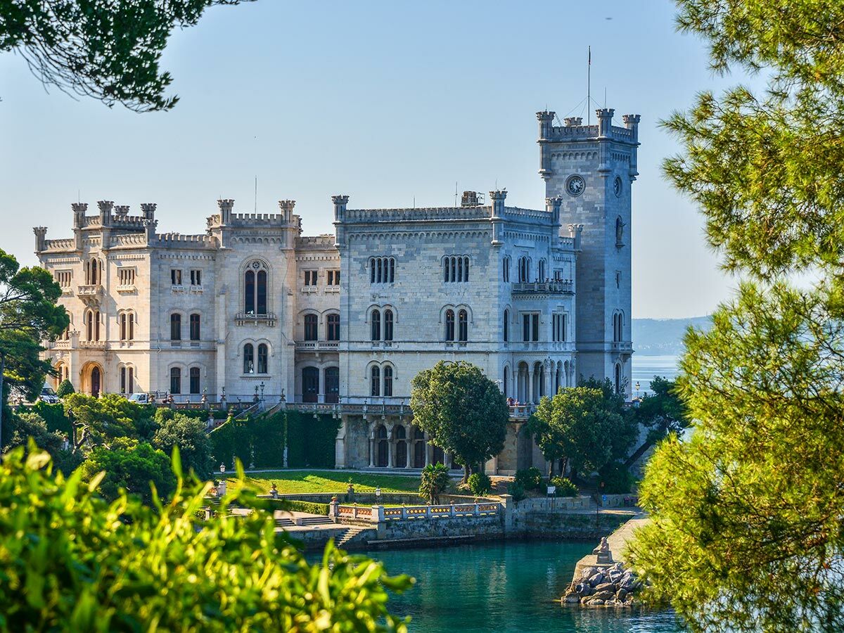 Historisches Schloss Miramare an der italienischen Adriaküste, umgeben von üppigem Grün und klarem Meer unter blauem Himmel.