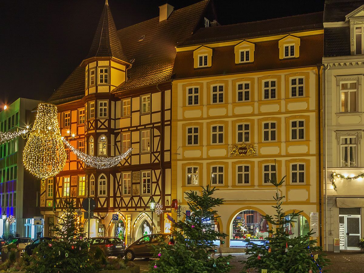 Abendliche Szene mit festlich beleuchteten historischen Gebäuden, Weihnachtsdekoration und kleinen Weihnachtsbäumen auf der Straße.