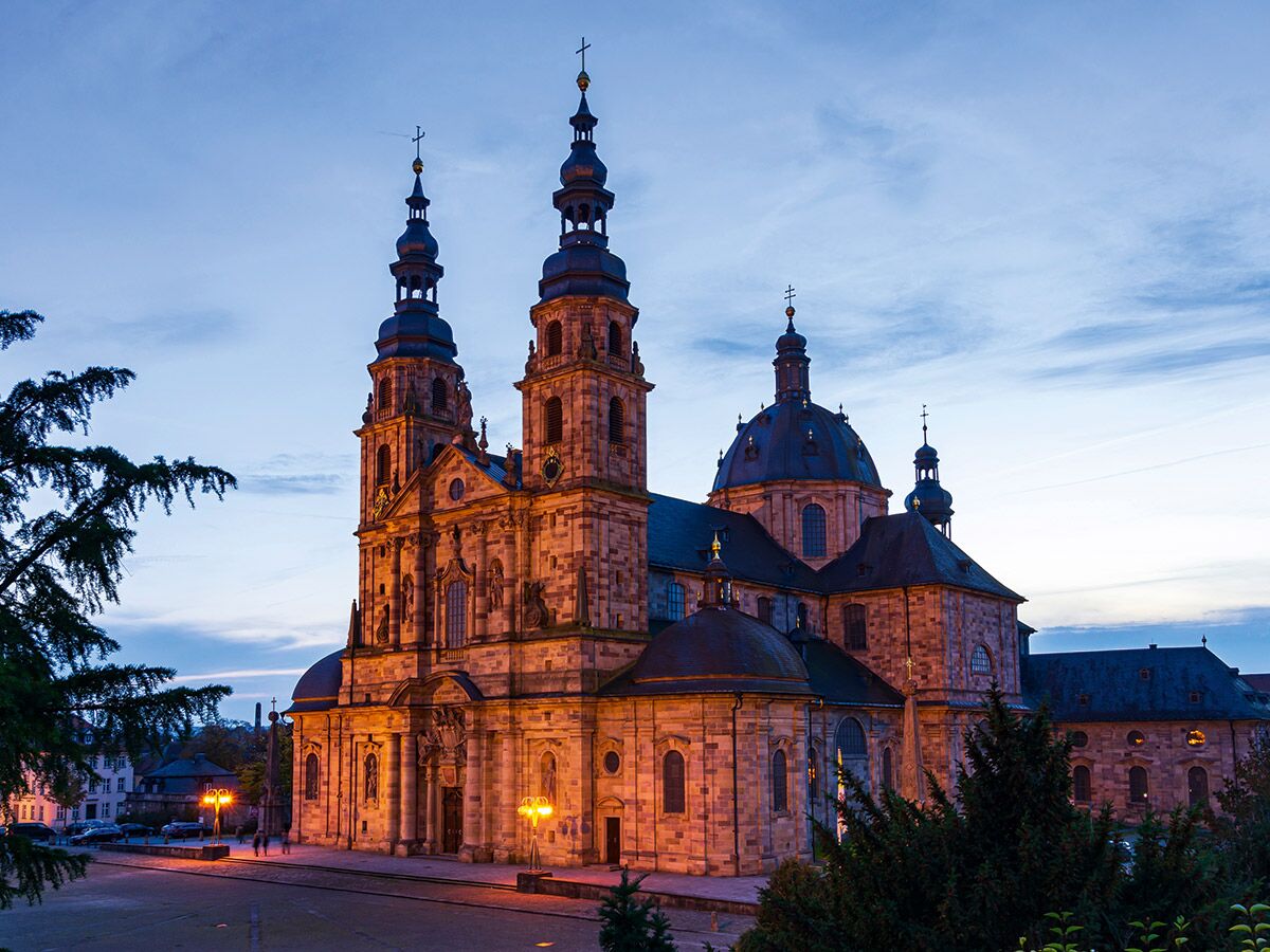 Die beleuchtete Fassade des Doms zu Fulda mit zwei Türmen und einer Kuppel bei Einbruch der Dämmerung.