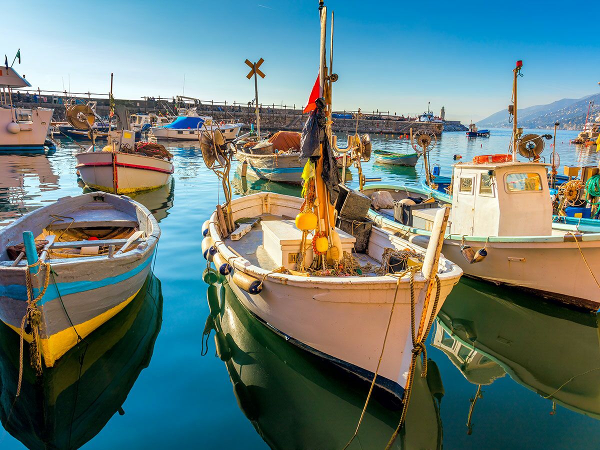 Bunte, traditionelle Fischerboote liegen friedlich in einem sonnigen Hafen mit blauem Wasser und klarem Himmel. Im Hintergrund sind Berge und ein Pier zu sehen.