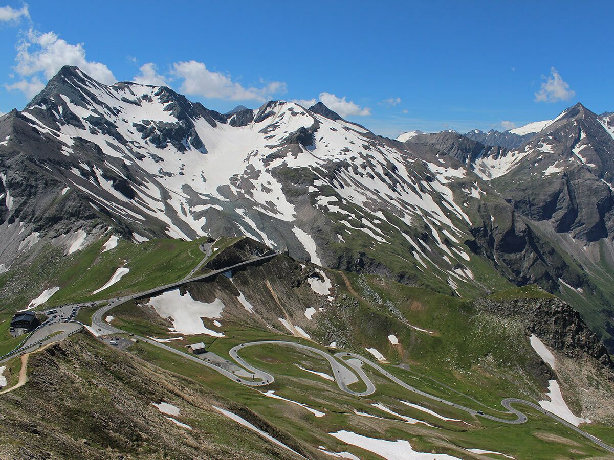 Panoramablick auf die Großglockner Hochalpenstraße mit Serpentinen, grünen Hängen, Schneebergen und blauem Himmel.