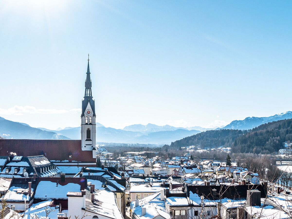 Panoramablick auf eine verschneite Alpenstadt mit markantem Kirchturm, umgeben von schneebedeckten Bergen unter blauem Himmel.