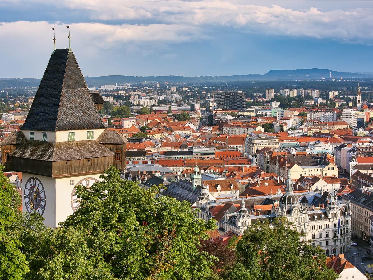 Panoramablick auf Graz mit dem berühmten Uhrturm im Vordergrund und der historischen Altstadt unter bewölktem Himmel.