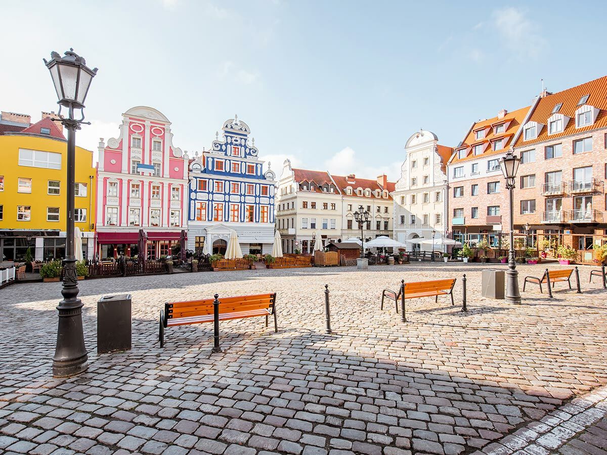 Malerischer Stadtplatz mit bunten historischen Gebäuden, Kopfsteinpflaster, Holzbänken und Laternen an einem sonnigen Tag.