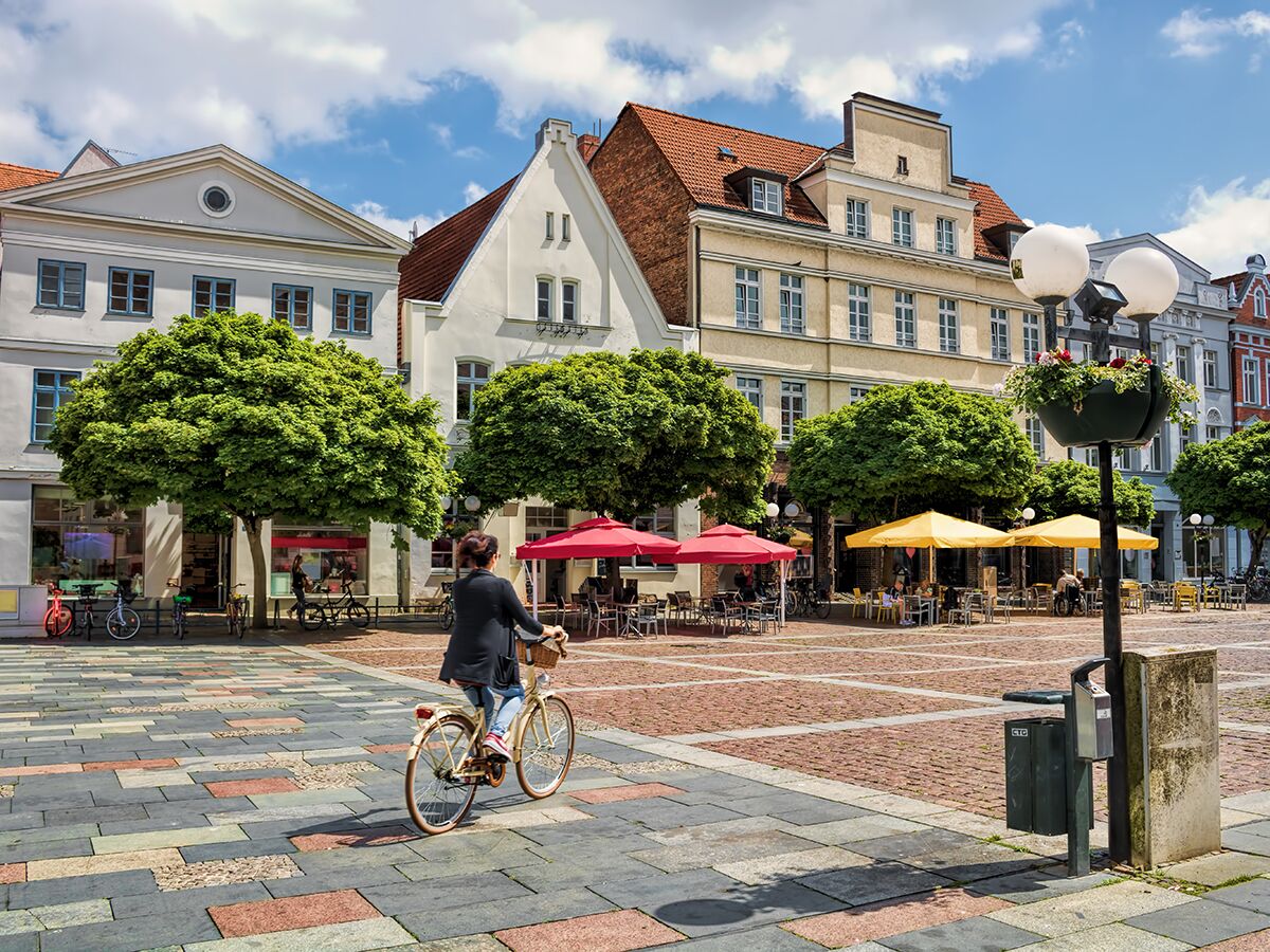 Frau fährt Rad auf belebtem historischen Marktplatz mit Cafés, Bäumen, Altbauten unter blauem Himmel in Deutschland.