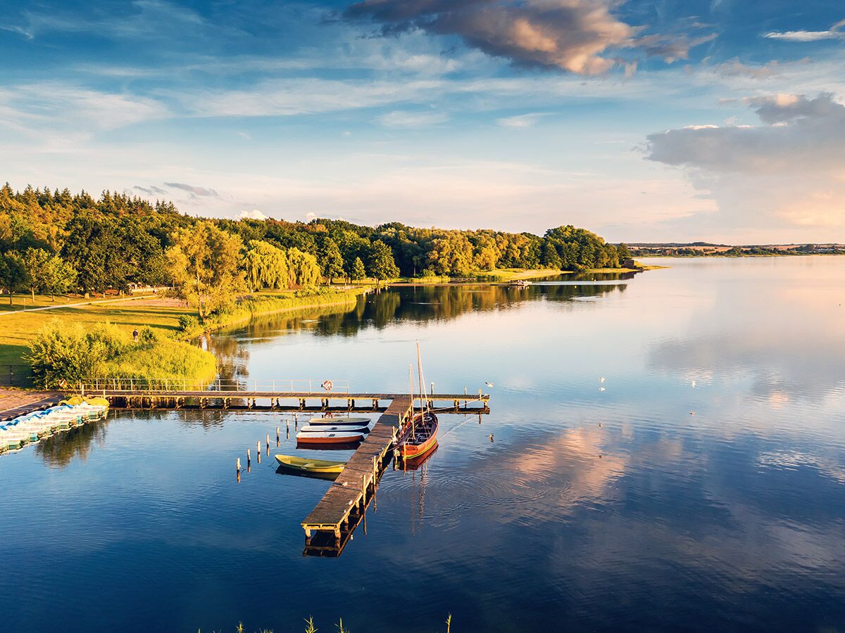 Ruhiger See mit Holzstegen, bunten Booten und grünen Bäumen am Ufer, beleuchtet vom warmen Licht des Sonnenuntergangs.