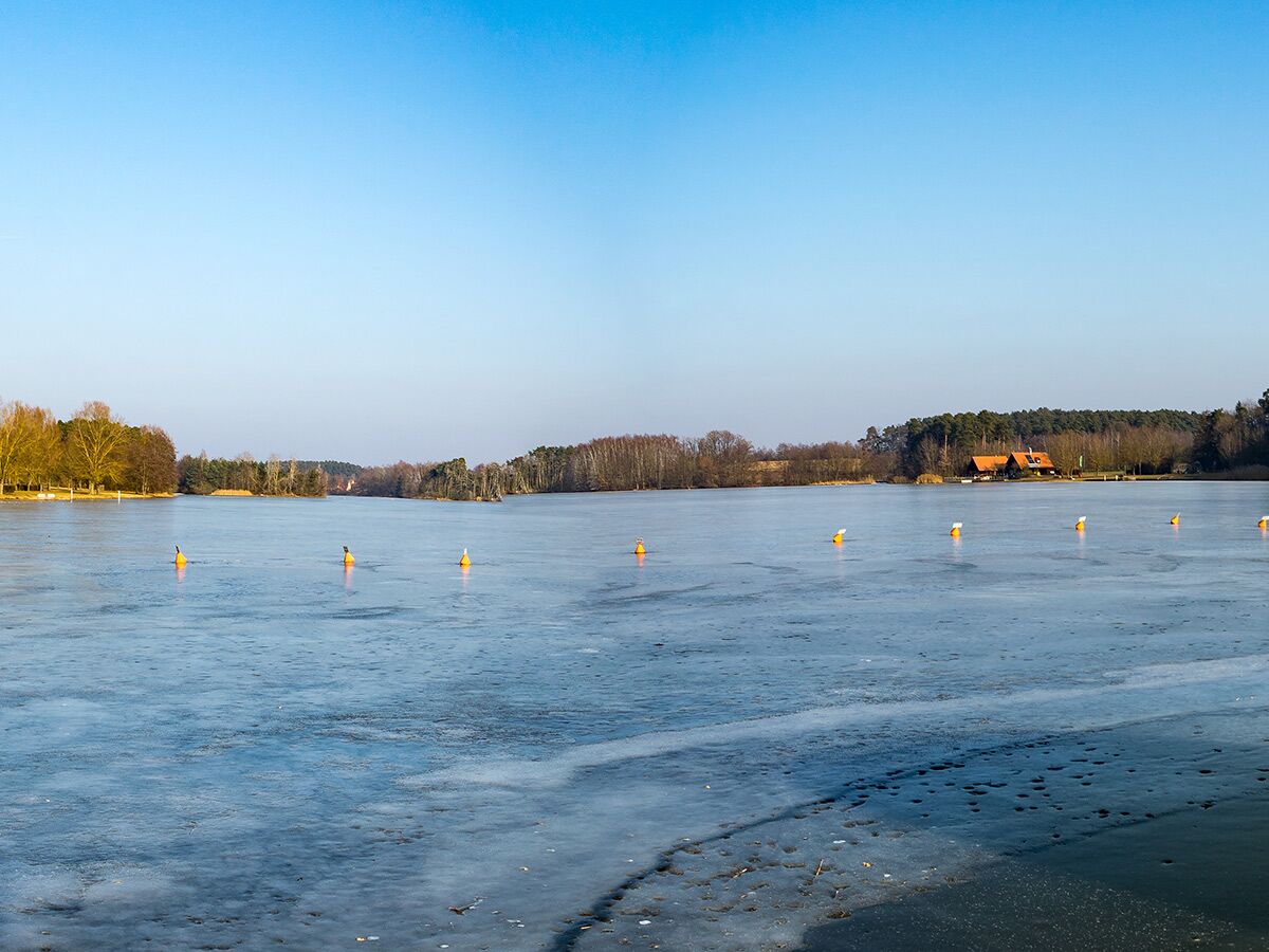 Weitblick auf einen teilweise gefrorenen See mit orangefarbenen Bojen, umgeben von Wald und einem Haus am Ufer unter blauem Himmel.
