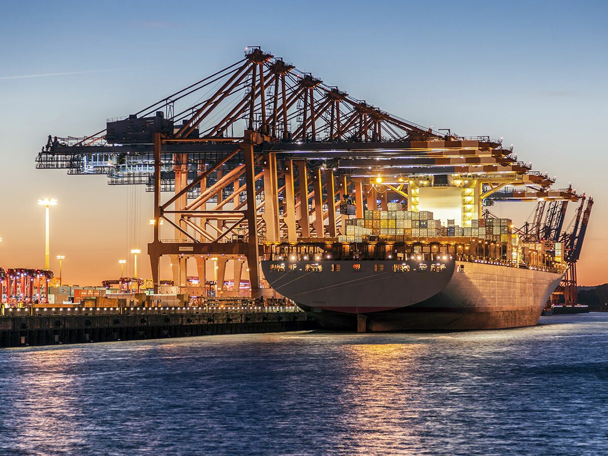 Großer Containerfrachter im beleuchteten Hafen bei Dämmerung, umgeben von riesigen Verladekränen.