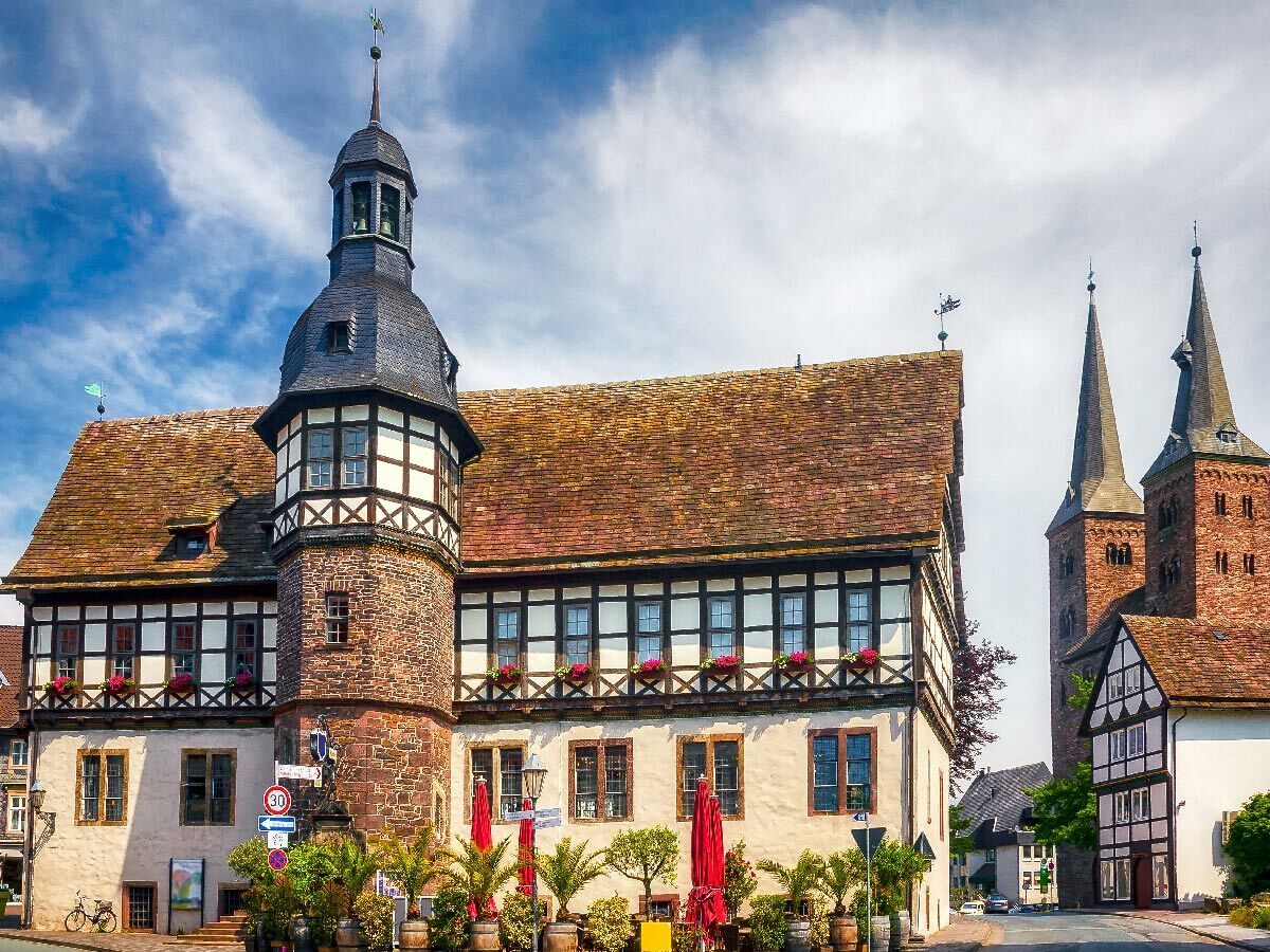 Historisches Fachwerkhaus mit Steinturm und Schieferdach, davor Pflanzen, im Hintergrund zwei Kirchtürme unter blauem Himmel.