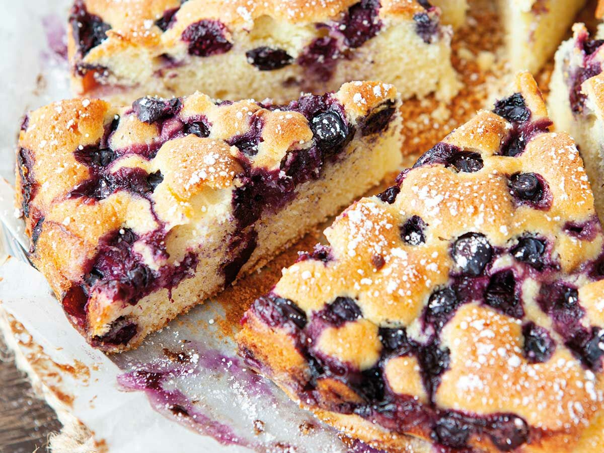 Nahaufnahme von frisch gebackenen Blaubeerkuchenstücken, großzügig mit Puderzucker bestreut, die lila Beeren sind gut sichtbar.