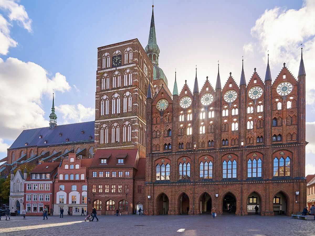 Historisches Rathaus Stralsund im Backsteingotik-Stil am belebten Marktplatz mit Nikolaikirche und blauem Himmel.