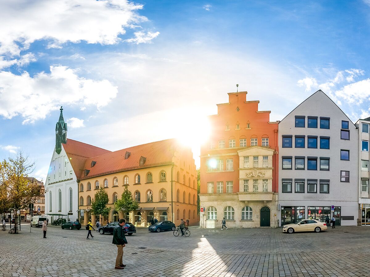 Panoramablick auf einen lebhaften deutschen Stadtplatz mit historischer Kirche, bunten Giebelhäusern und Menschen an einem sonnigen Tag.