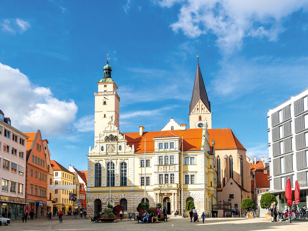 Historischer Marienplatz in Landsberg am Lech mit Altem Rathaus, Stadtpfarrkirche und belebtem Stadtleben unter blauem Himmel.