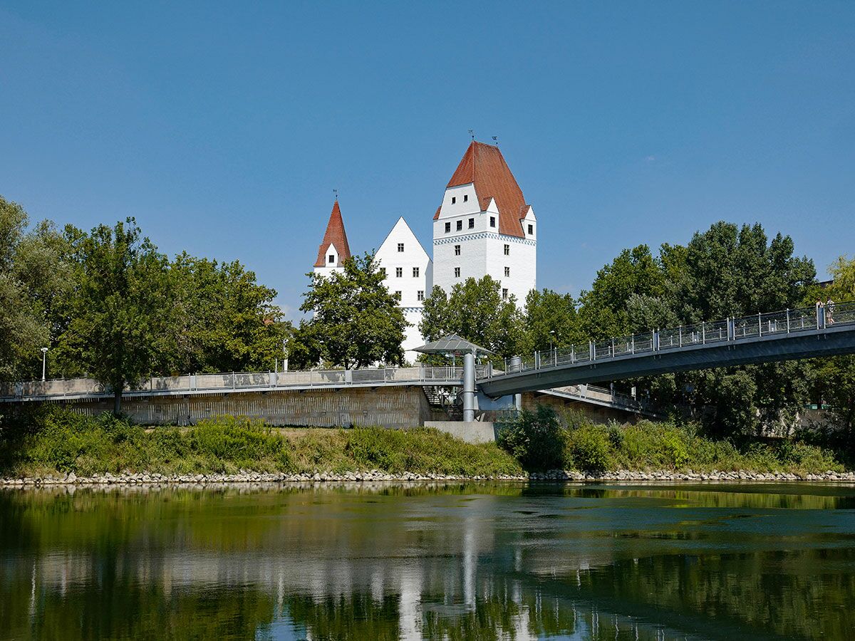 Burg Trausnitz in Landshut an der Isar mit rotem Dach, umgeben von Bäumen und einer modernen Fußgängerbrücke. Sonniger Tag.