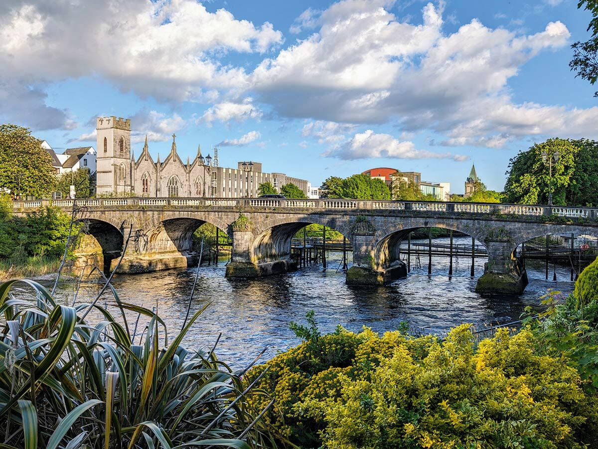 Historische Steinbrücke über den Fluss Corrib in Galway, Irland, mit der Galway Cathedral im Hintergrund unter blauem Himmel.