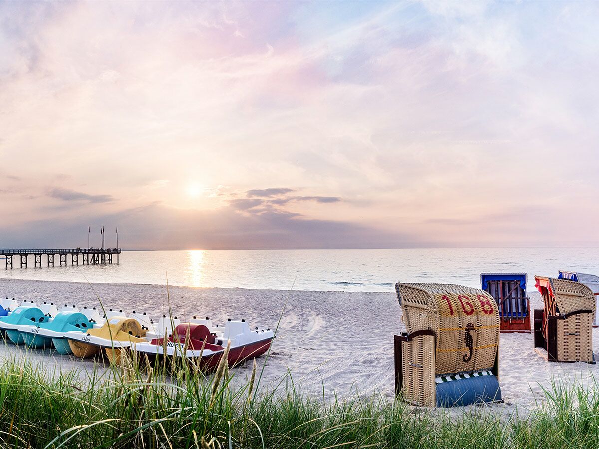 Idyllischer Sonnenuntergang am Sandstrand mit bunten Tretbooten, Strandkörben und einer Seebrücke im Hintergrund.