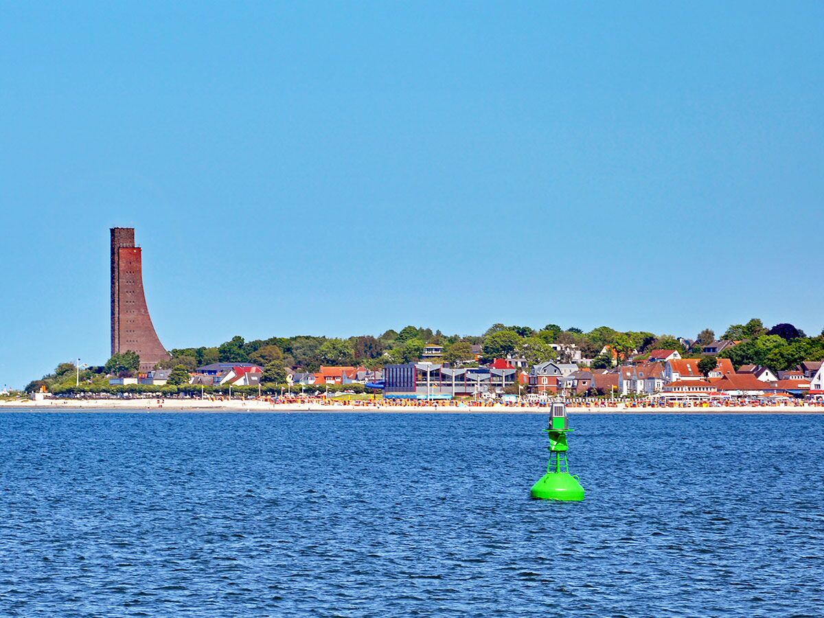 Panoramablick auf das Marine-Ehrenmal Laboe, den sonnigen Strand mit Badegästen und die Ostsee mit einer grünen Boje.