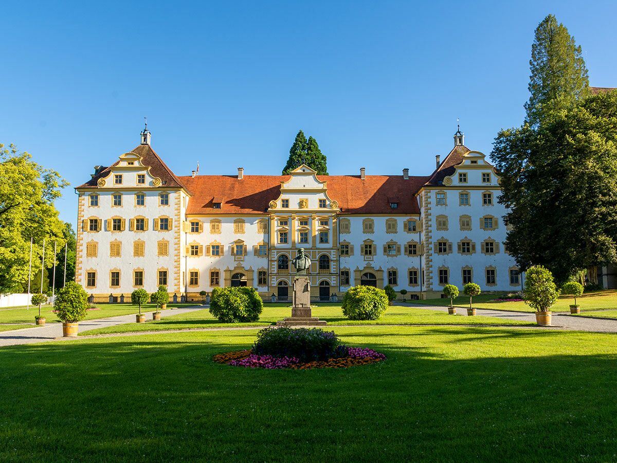 Weißes historisches Schloss im Barockstil mit roten Dächern, gelben Fensterläden und einer Statue in einem grünen Park unter blauem Himmel.