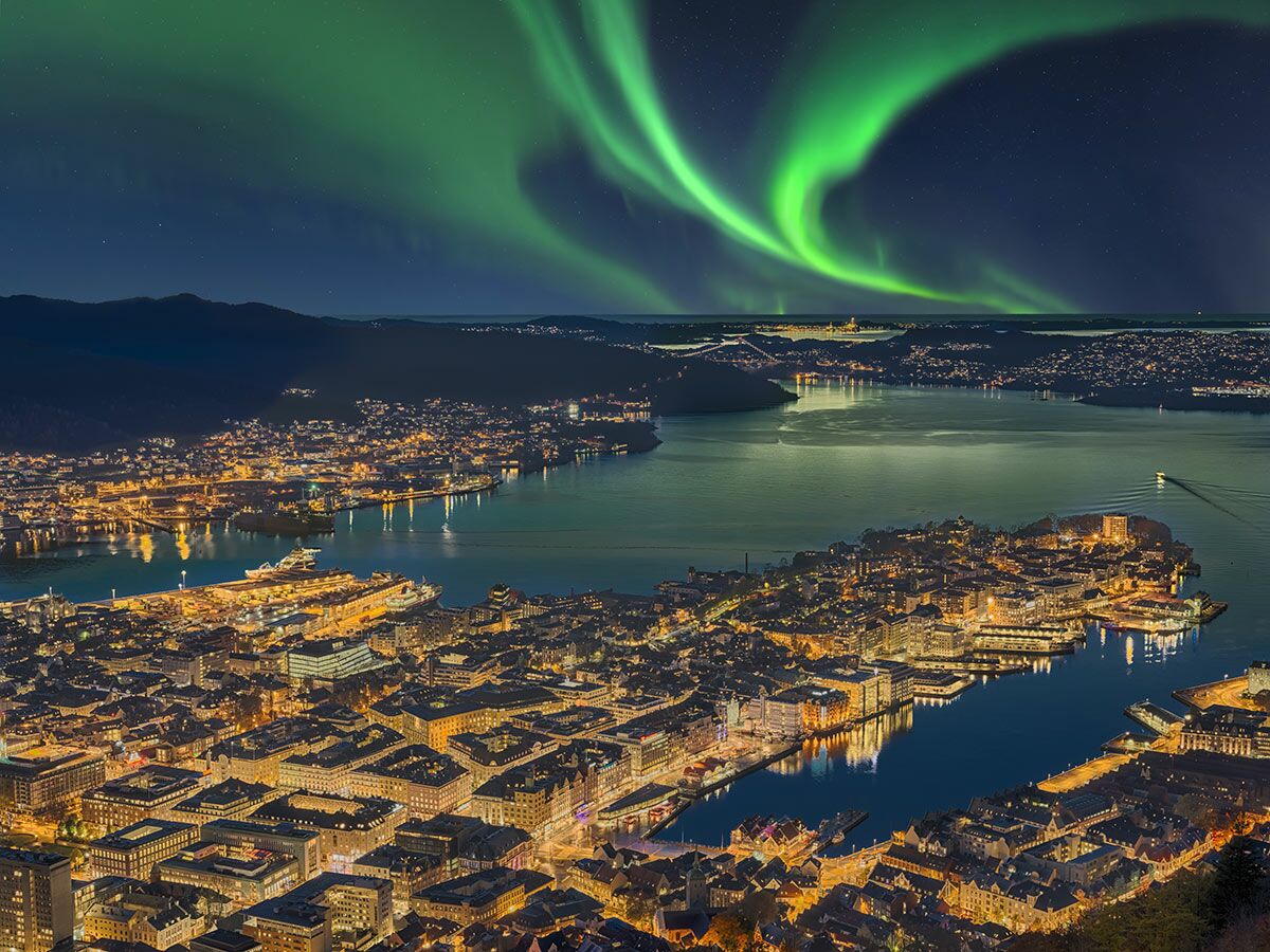 Nordlichter tanzen über Bergen, Norwegen. Eine beleuchtete Stadt bei Nacht mit Hafen, Bergen und Fjorden unter einem Sternenhimmel.