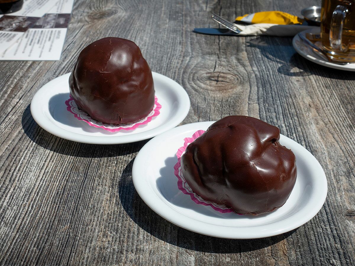 Zwei glänzende Schokoladen-Gebäcke auf weißen Tellern mit rosa Papieruntersetzern auf rustikalem Holztisch im Sonnenlicht.
