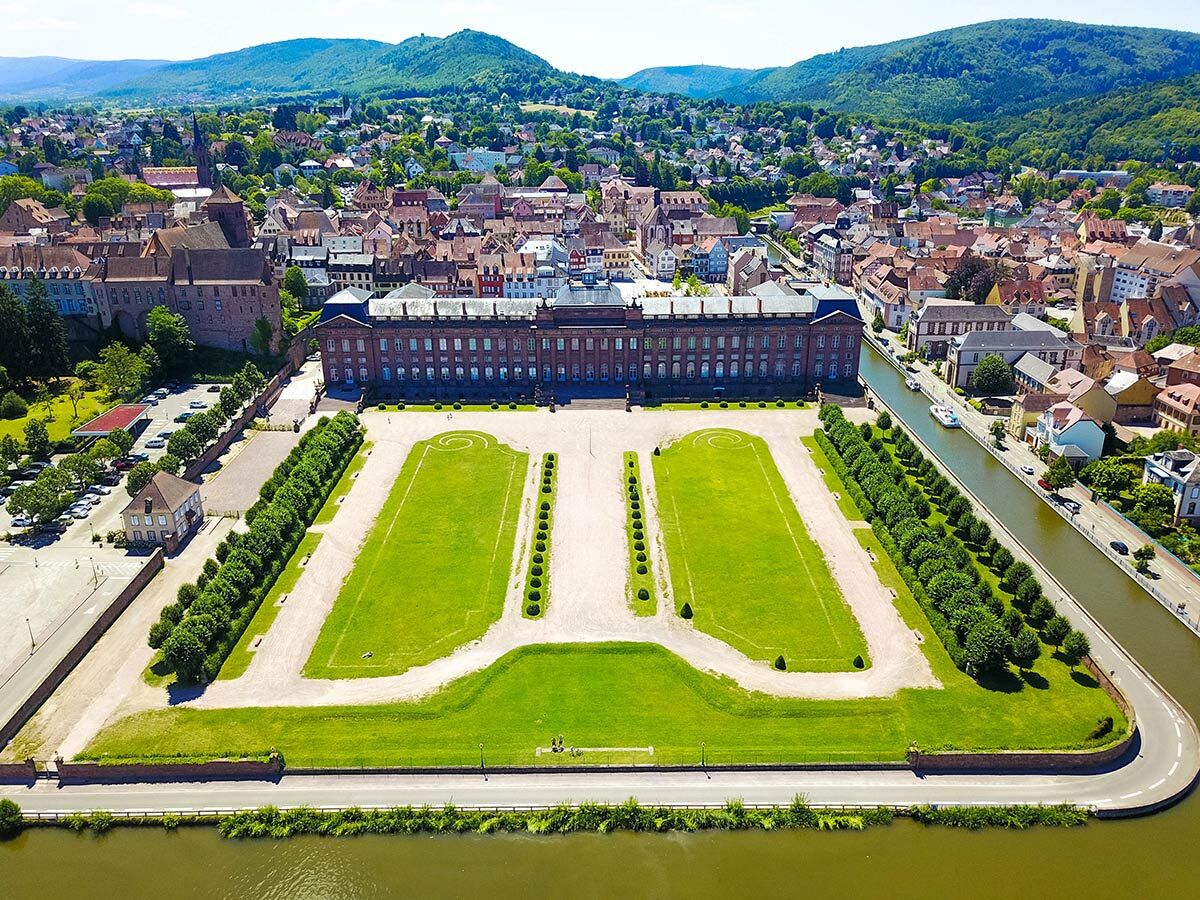Luftaufnahme des Schlosses Lunéville mit seinen symmetrischen Gärten, umgeben von einer historischen Stadt und einem Fluss unter grünen Hügeln.