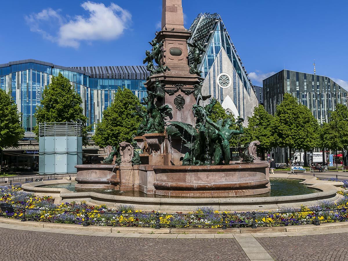 Ornate Mendebrunnen Springbrunnen mit Skulpturen vor modernen Gebäuden und Paulinum in Leipzig bei blauem Himmel.
