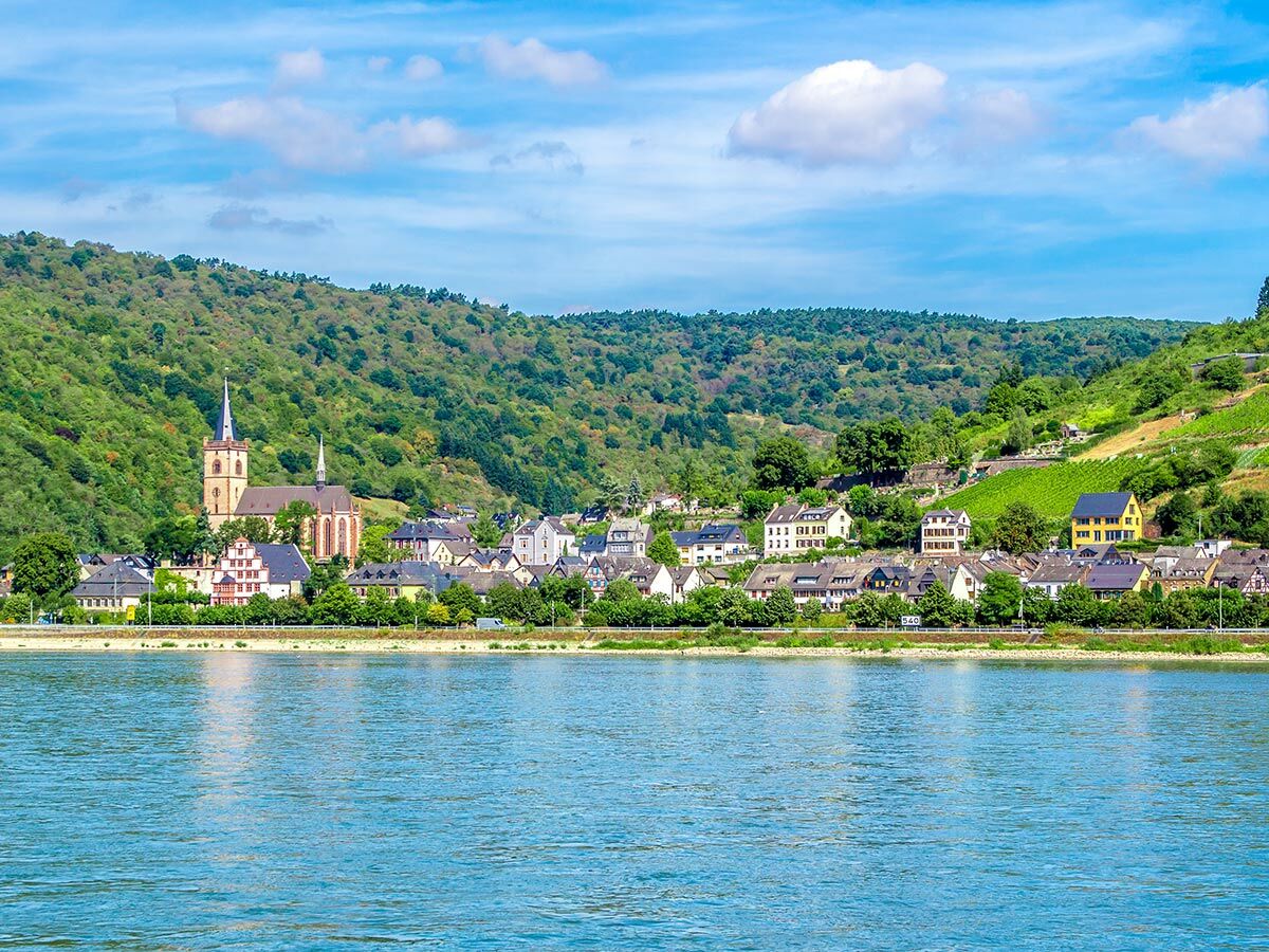 Malerisches Dorf am Rhein mit historischer Kirche, grünen Hügeln und Weinbergen unter blauem Himmel.