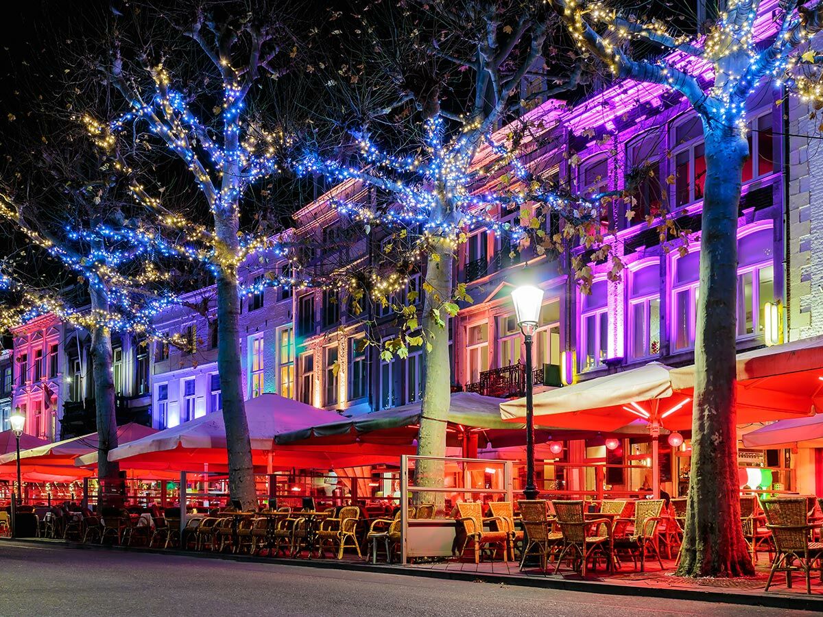 Beleuchtete Stadtstraße bei Nacht mit Außencafés, roten Markisen, Tischen und Stühlen unter Bäumen mit blauen und gelben Lichterketten.