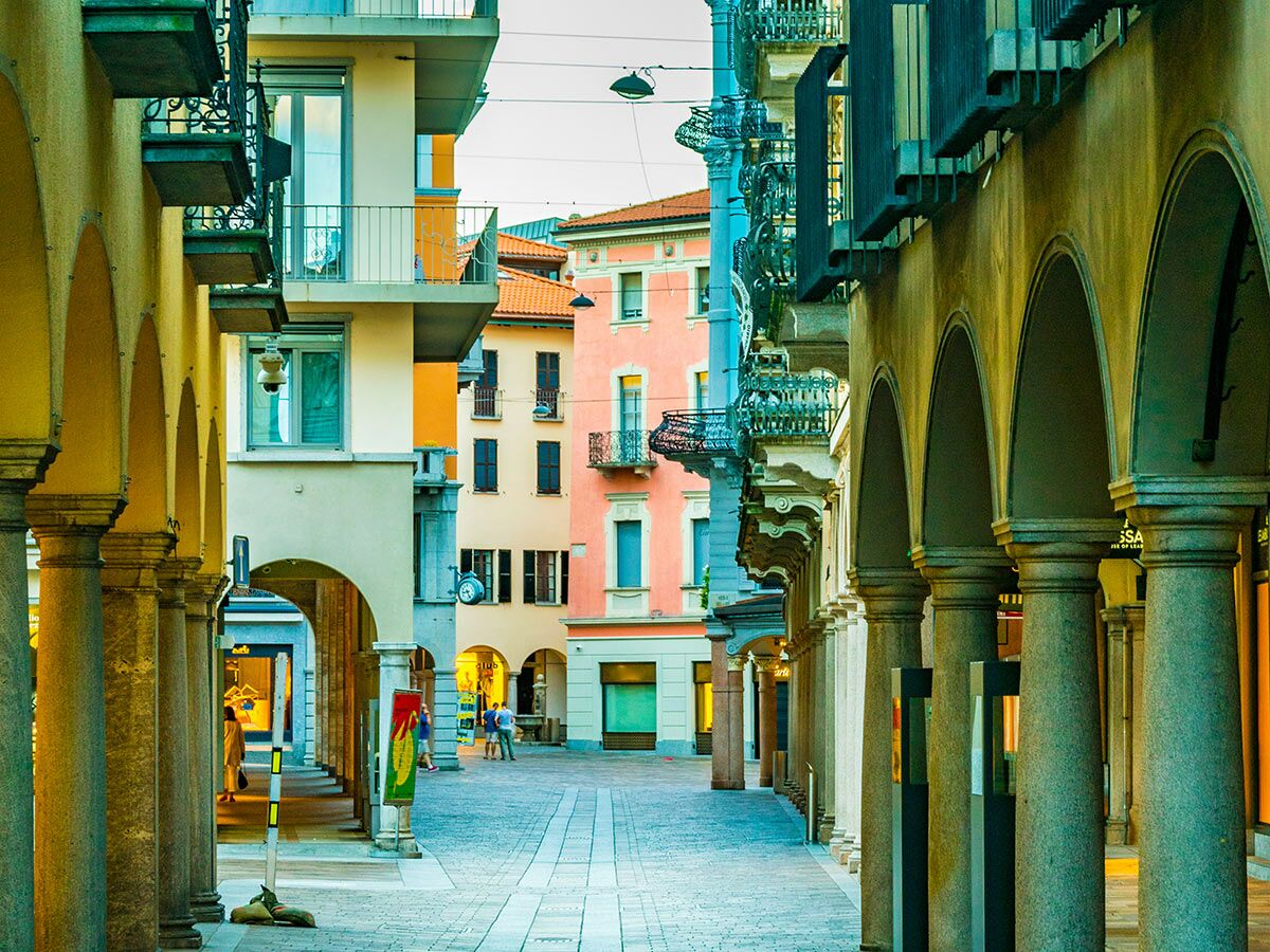 Arkadengasse mit bunten Häusern und Passanten in einer südeuropäischen Stadt. Historische Architektur prägt das Bild.