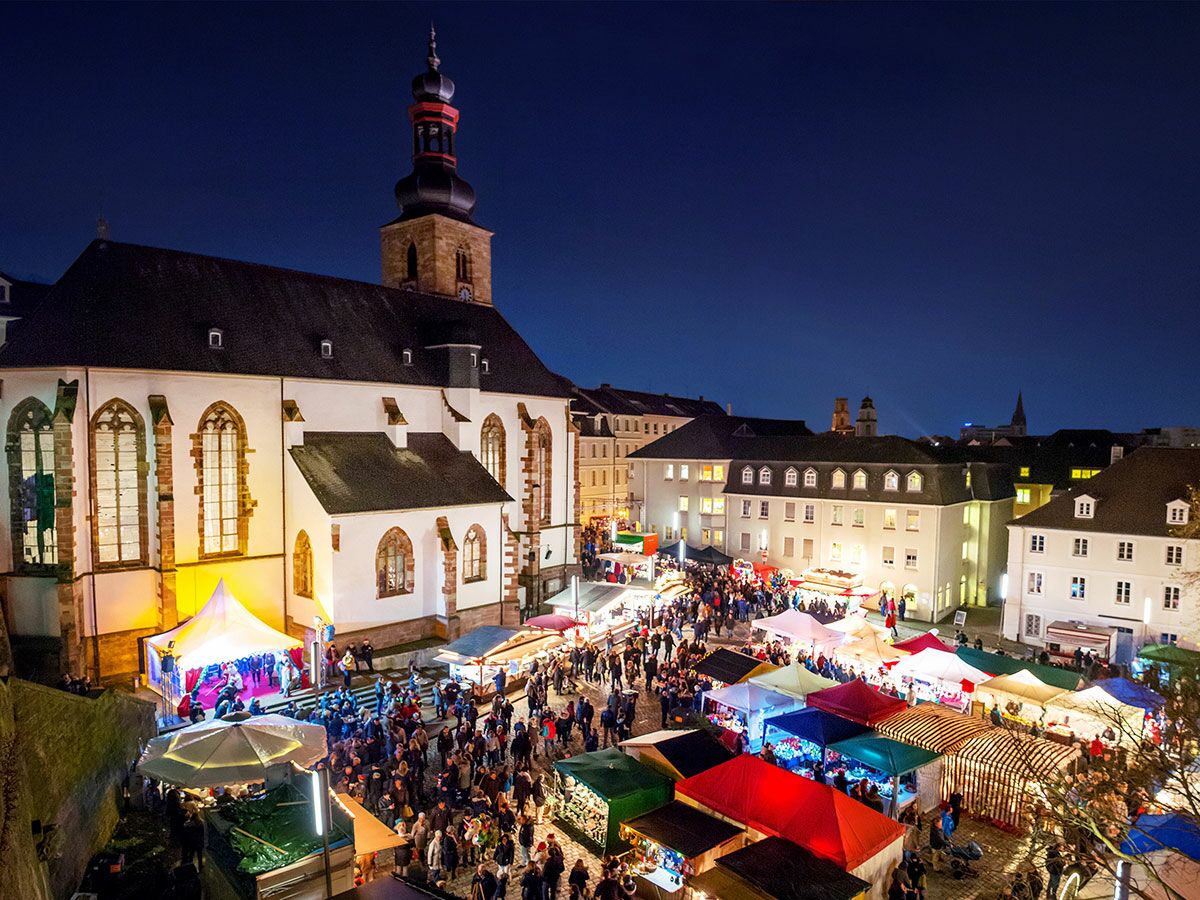 Abendliche Luftaufnahme eines belebten Weihnachtsmarktes mit bunten Ständen, vielen Menschen und einer großen Kirche im Hintergrund.
