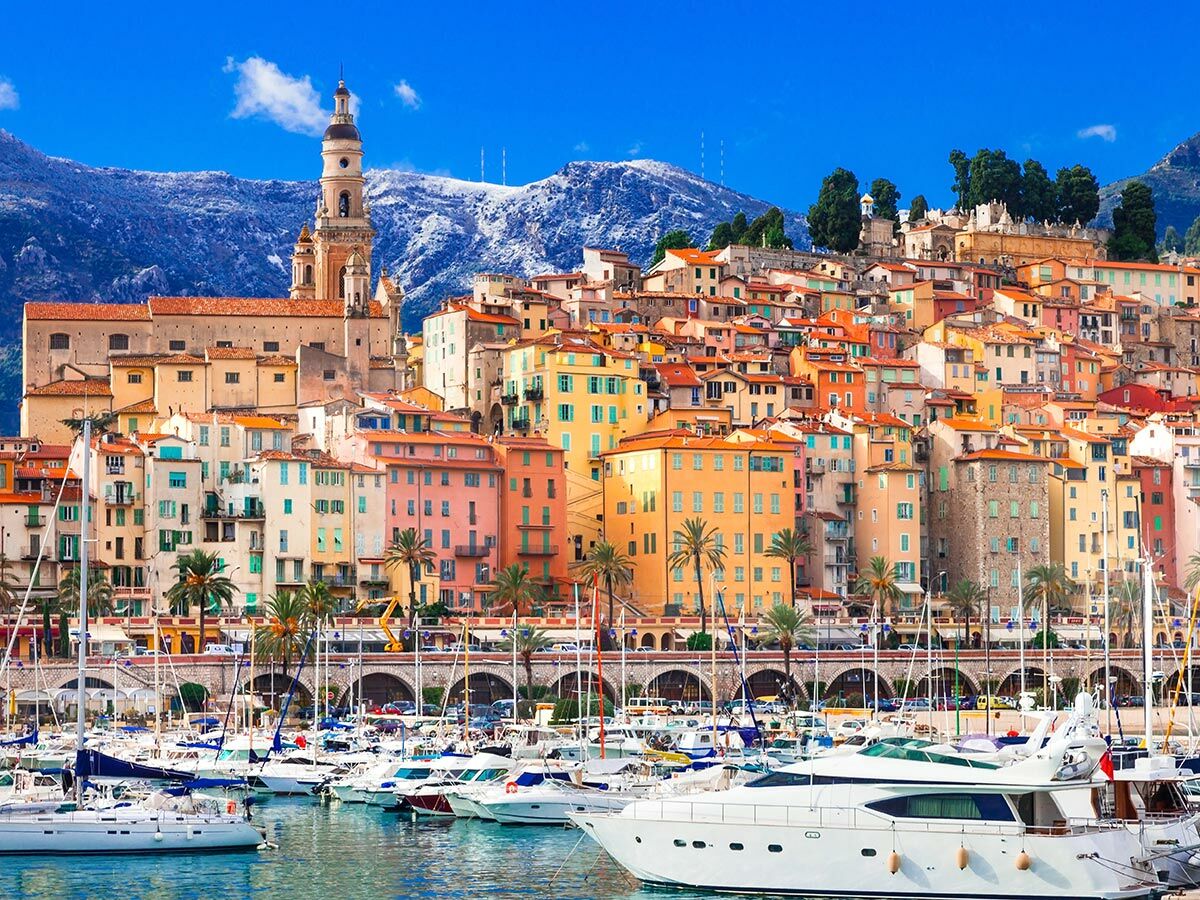 Panoramablick auf Menton mit bunten Häusern der Altstadt, Yachthafen und schneebedeckten Bergen unter blauem Himmel.