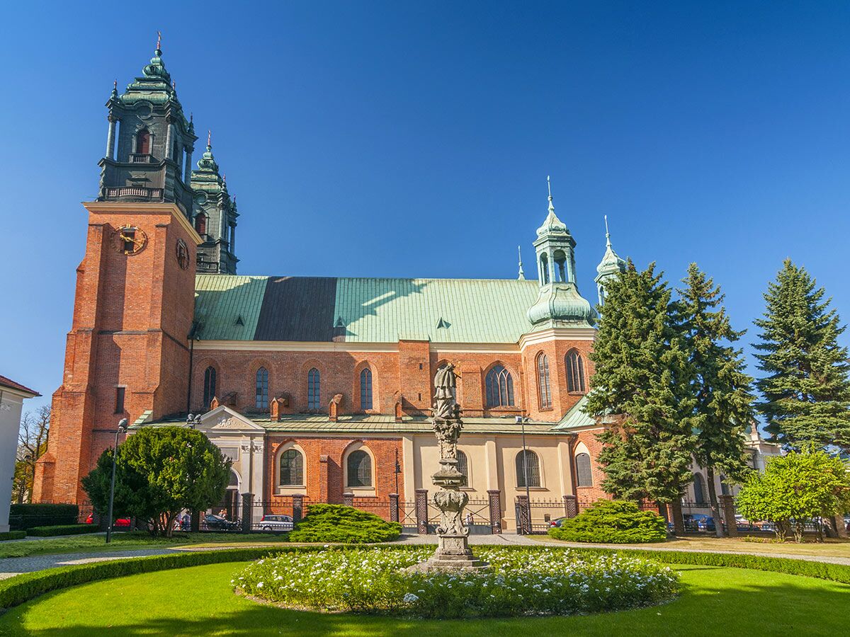 Historischer Gnesener Dom, Polen, mit roten Ziegelmauern, grünen Dächern, barocken Türmen und einer Statue unter blauem Himmel.